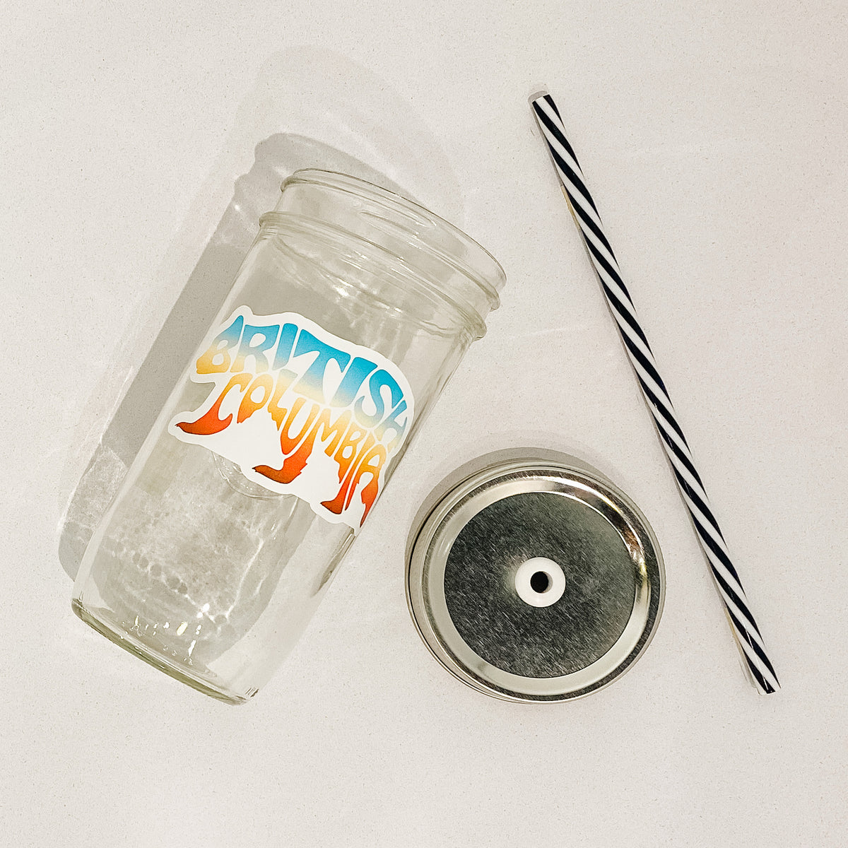 Flashy lay of mason jar tumbler that reads British Columbia in shape of bear. Colour is bright sunset gradient. Black and white striped straw. Mason jar straw lid is plain silver.