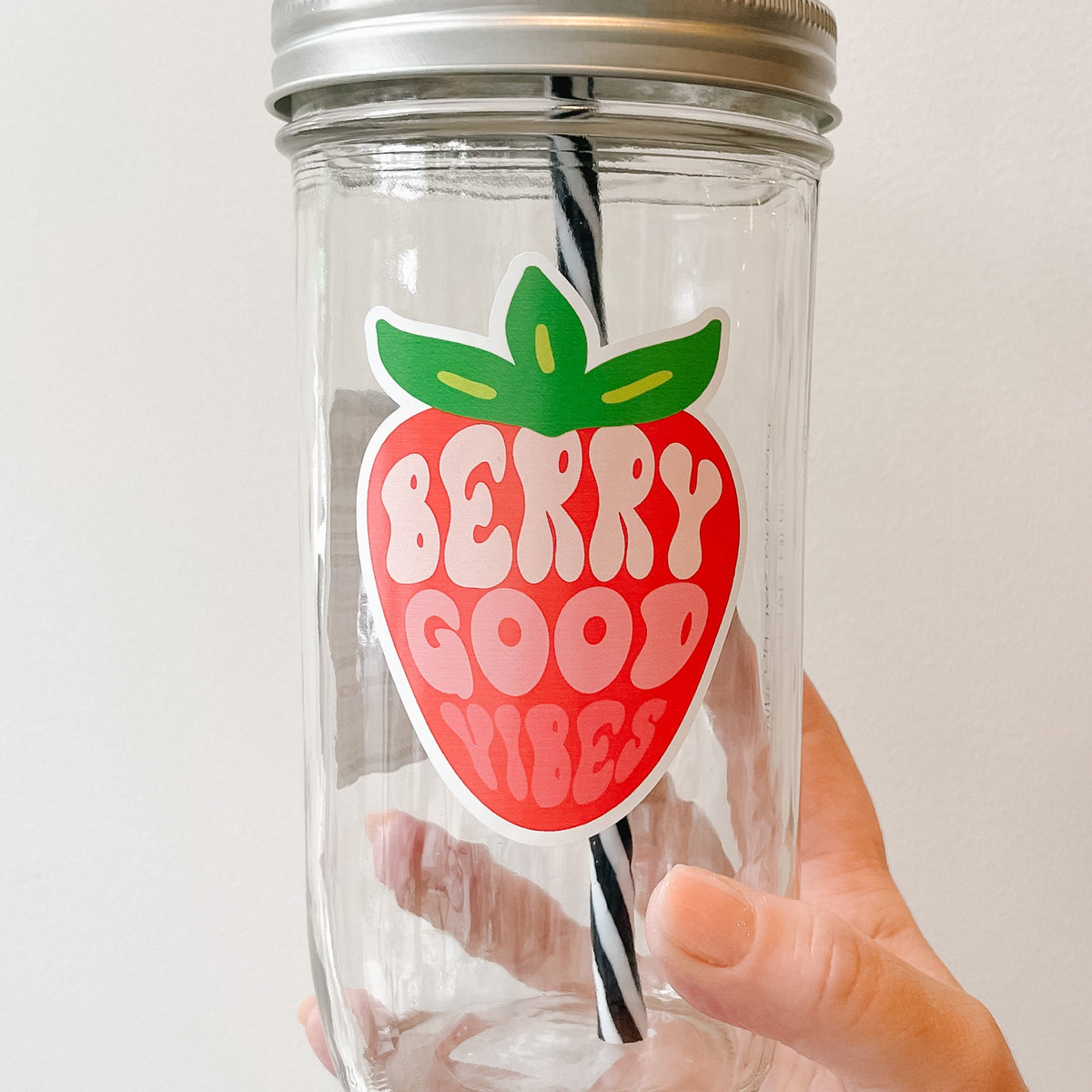 Close up of a hand holding a mason jar tumbler that has a strawberry on it with the words “berry good vibes” within the strawberry in different shades of pink to depict the colours within an actual strawberry.