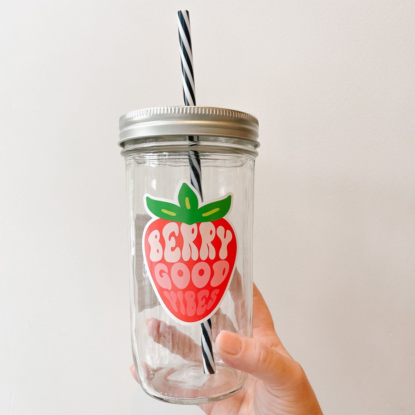 Hand holding a mason jar tumbler that has a strawberry on it with the words “berry good vibes” within the strawberry in different shades of pink to depict the colours within an actual strawberry.