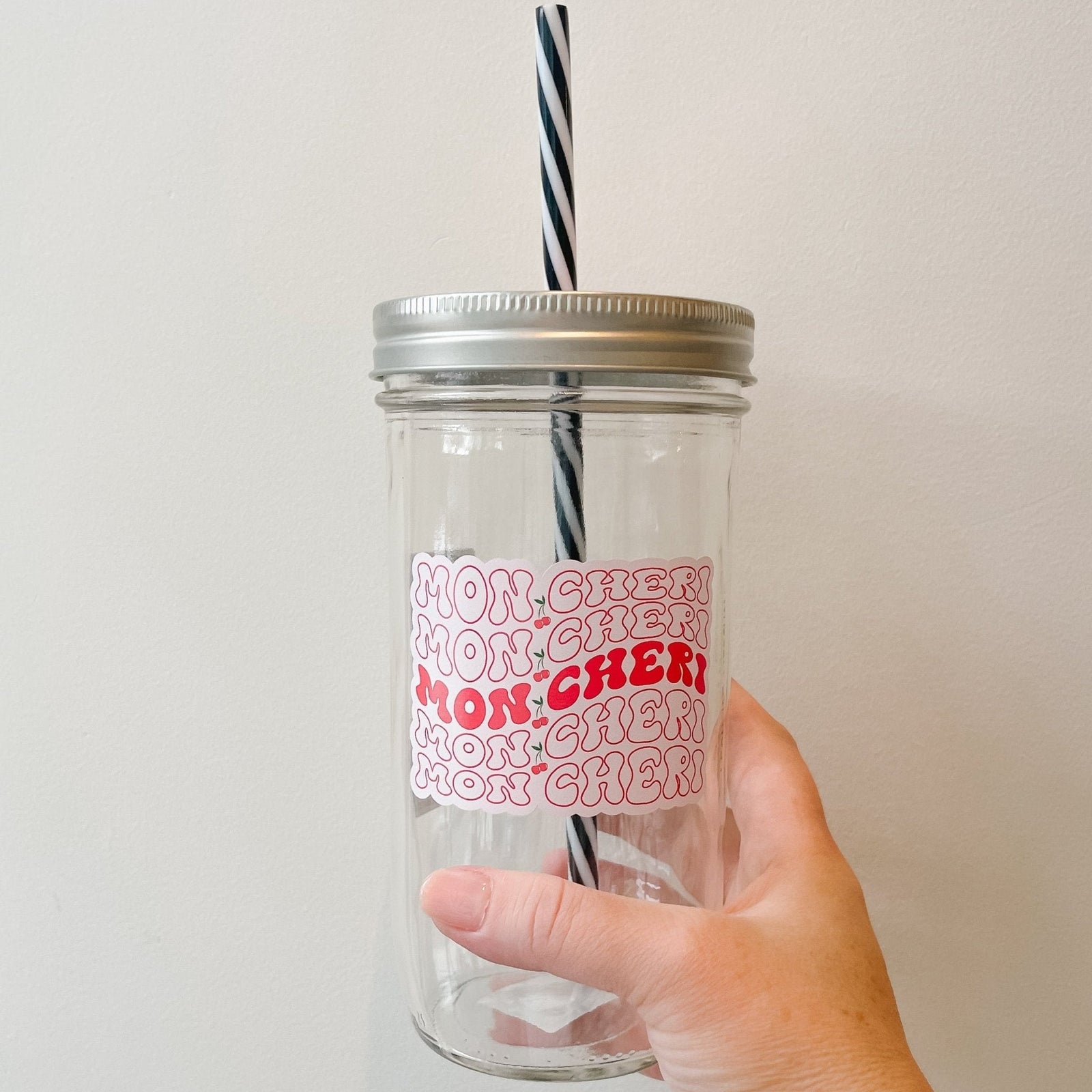 Hand holding a mason jar tumbler that says “mon cheri” with a cherry in between the words. This repeats 5 times, the words are outlined red except for the middle one where the words are filled in red. All on a pink background.