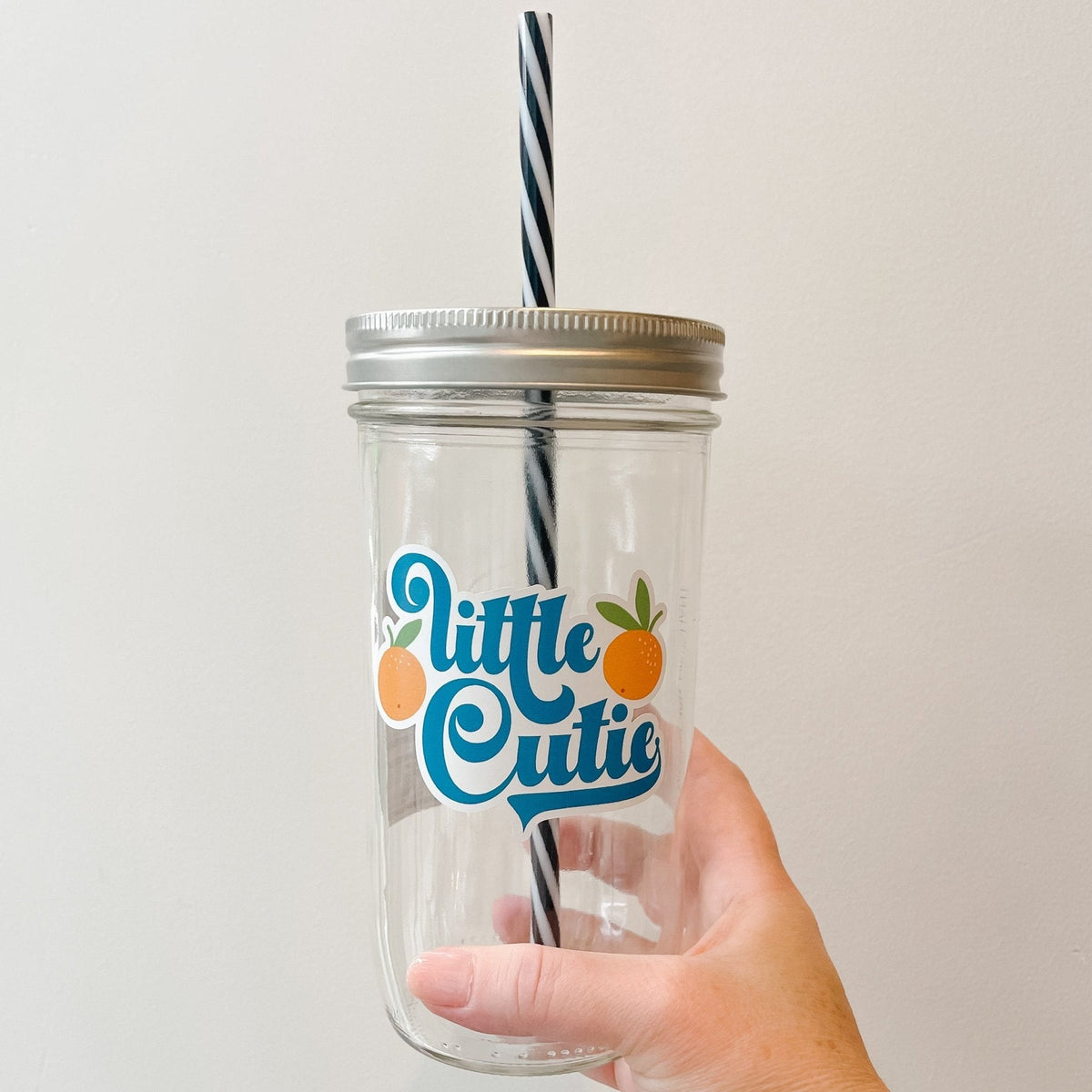 Hand holding a mason jar tumbler that says “little cutie” with two oranges on either side of the words.