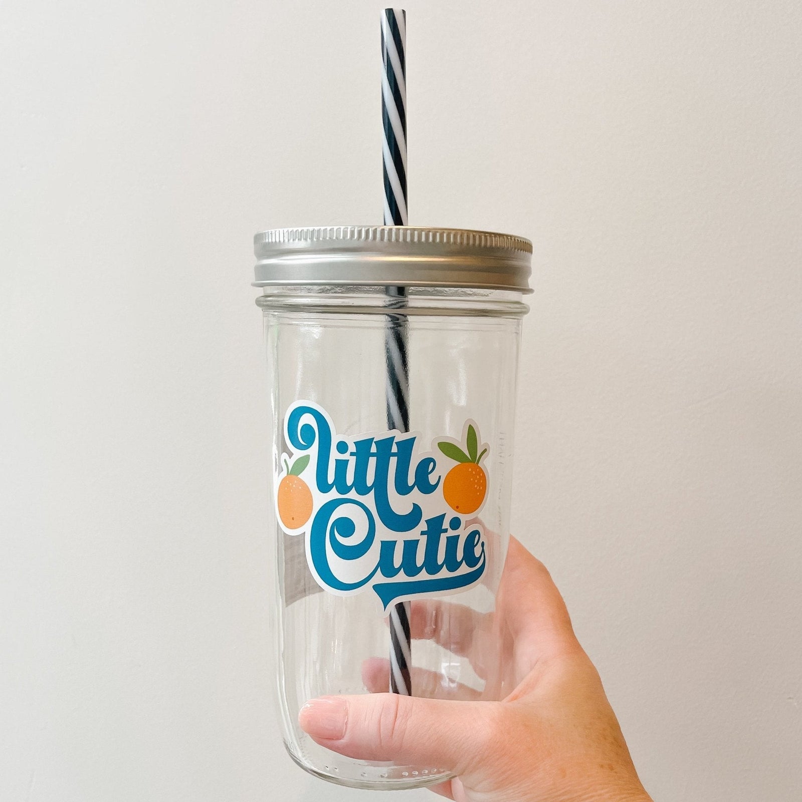 Hand holding a mason jar tumbler that says “little cutie” with two oranges on either side of the words.