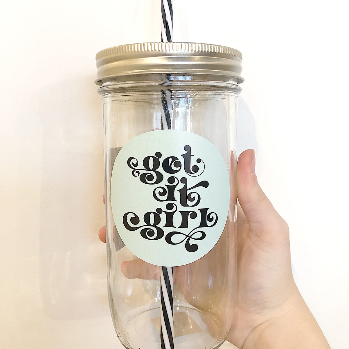 Hand holding a mason jar tumbler that says “get it girl” in black on a light mint circular background.