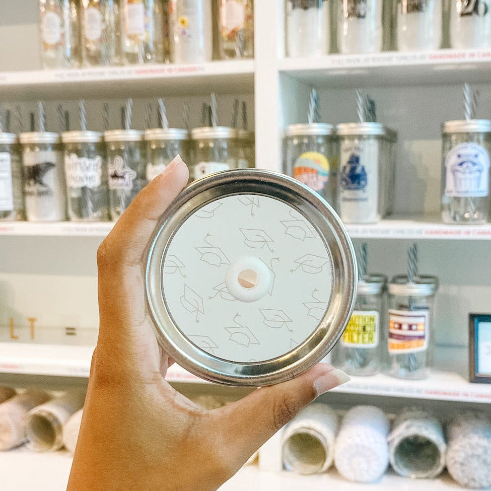 Hand holding mason jar patterned straw lid that has gold grad caps on white background