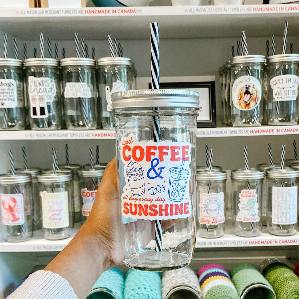 Hand holding a mason jar tumbler that reads iced coffee and sunshine all day every day in red ink with drink designs and ice cubes on white background