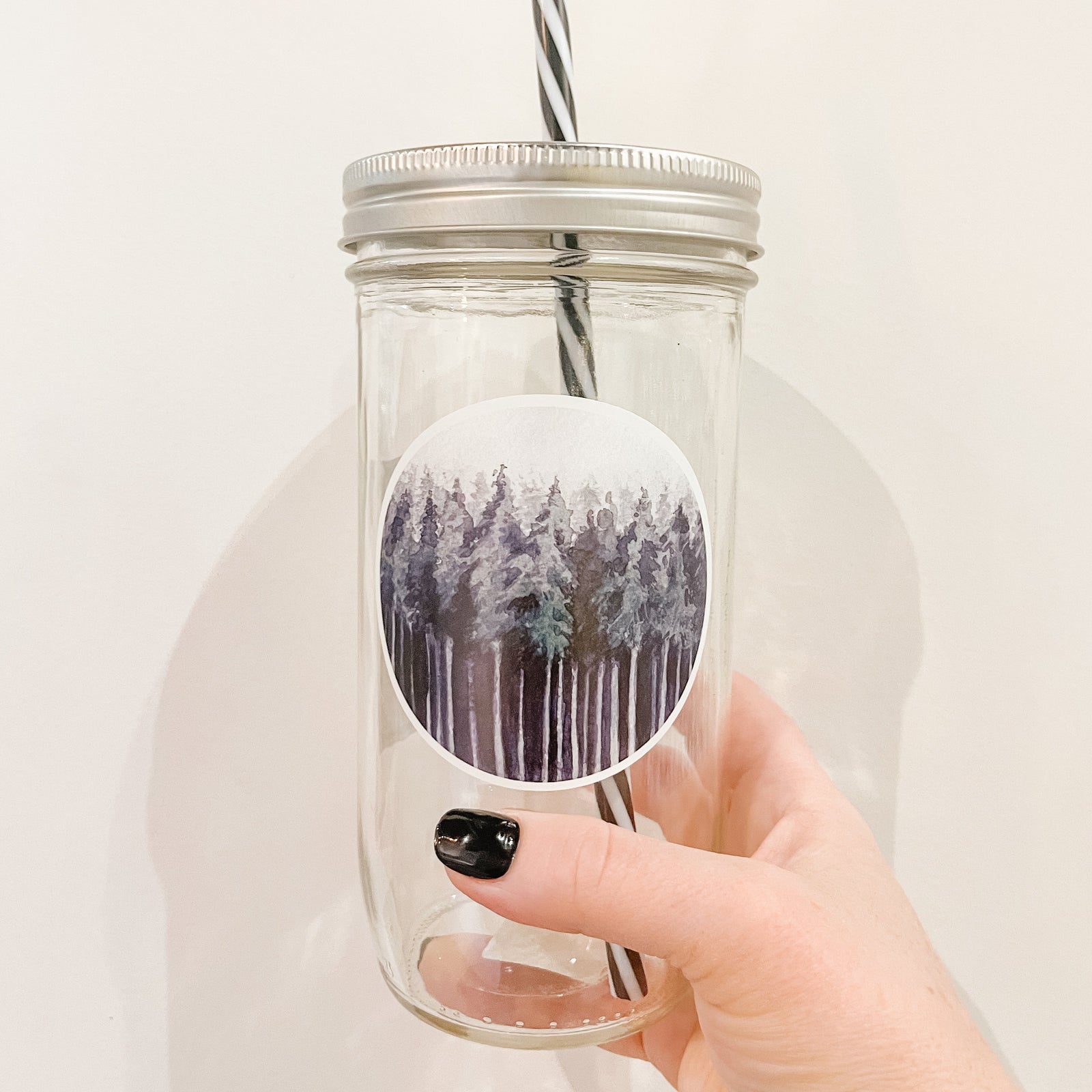 Hand holding a mason jar tumbler with a circular decal of multiple trees in shades of dark green and greys.