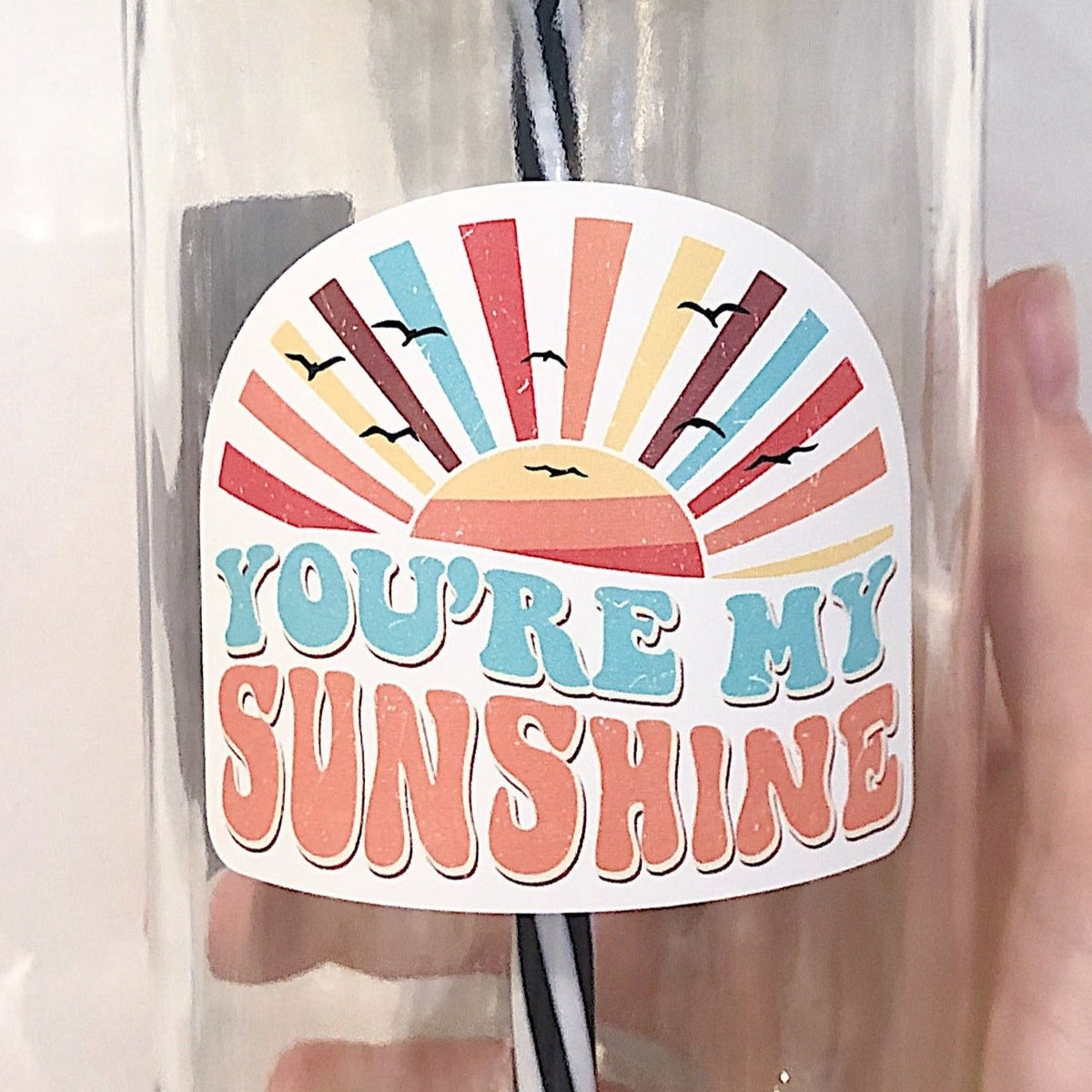 Close up of a hand holding a mason jar tumbler that has an image of a multicoloured sunset and says "you're my sunshine"