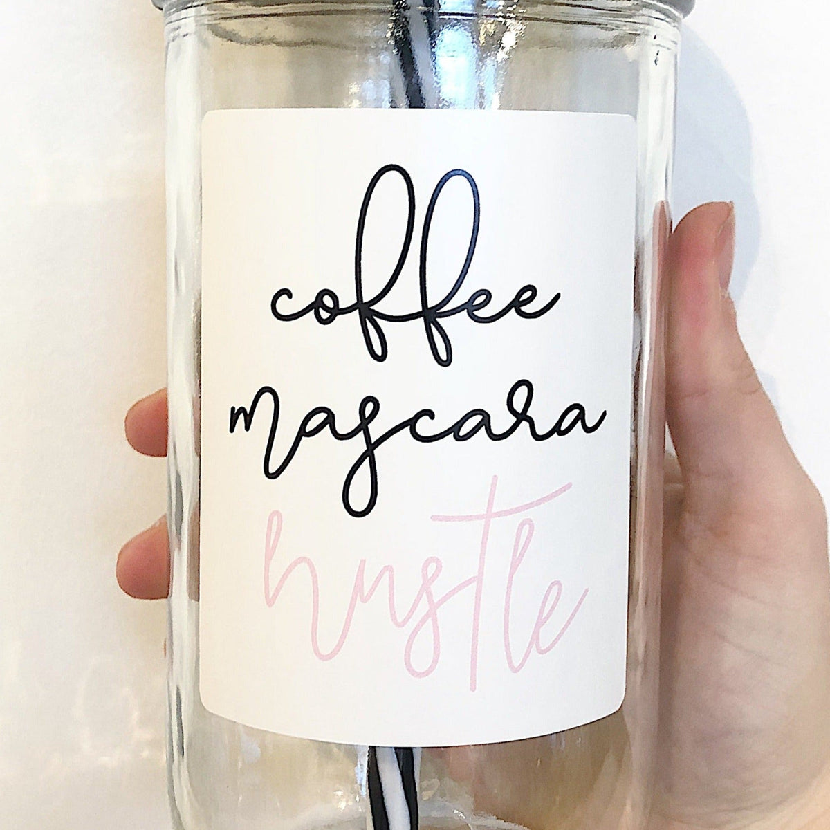 Close up of a hand holding a mason jar tumbler that says “coffee mascara hustle”.