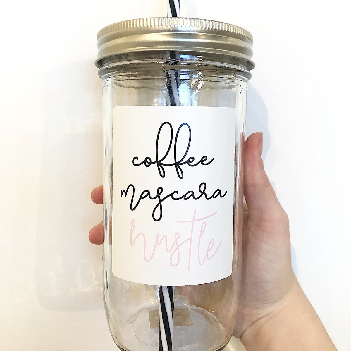 Hand holding a mason jar tumbler that says “coffee mascara hustle” with coffee and mascara in black and hustle in pink on a white background.