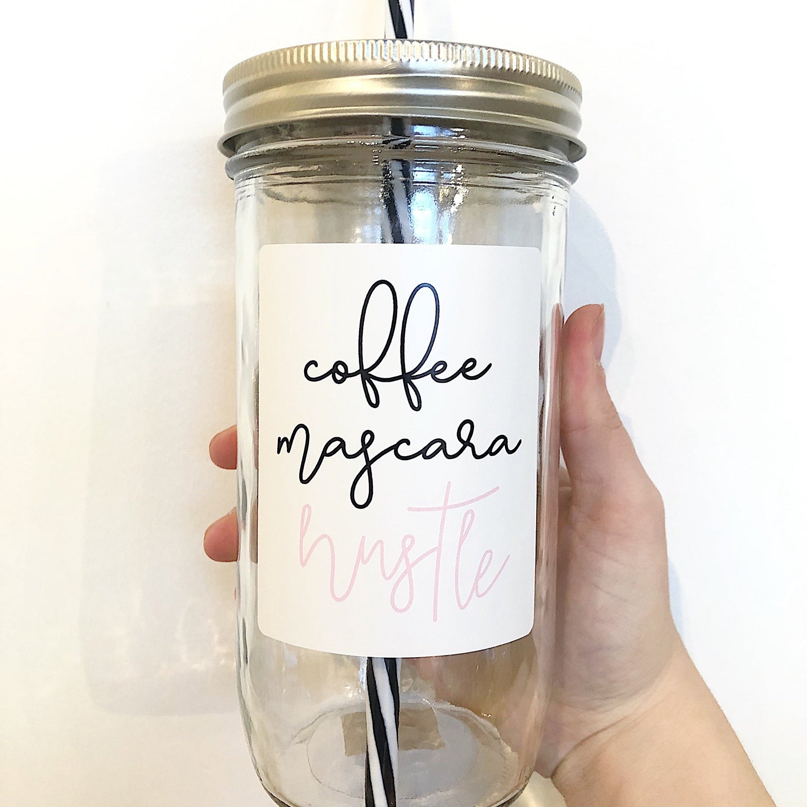Hand holding a mason jar tumbler that says “coffee mascara hustle” with coffee and mascara in black and hustle in pink on a white background.