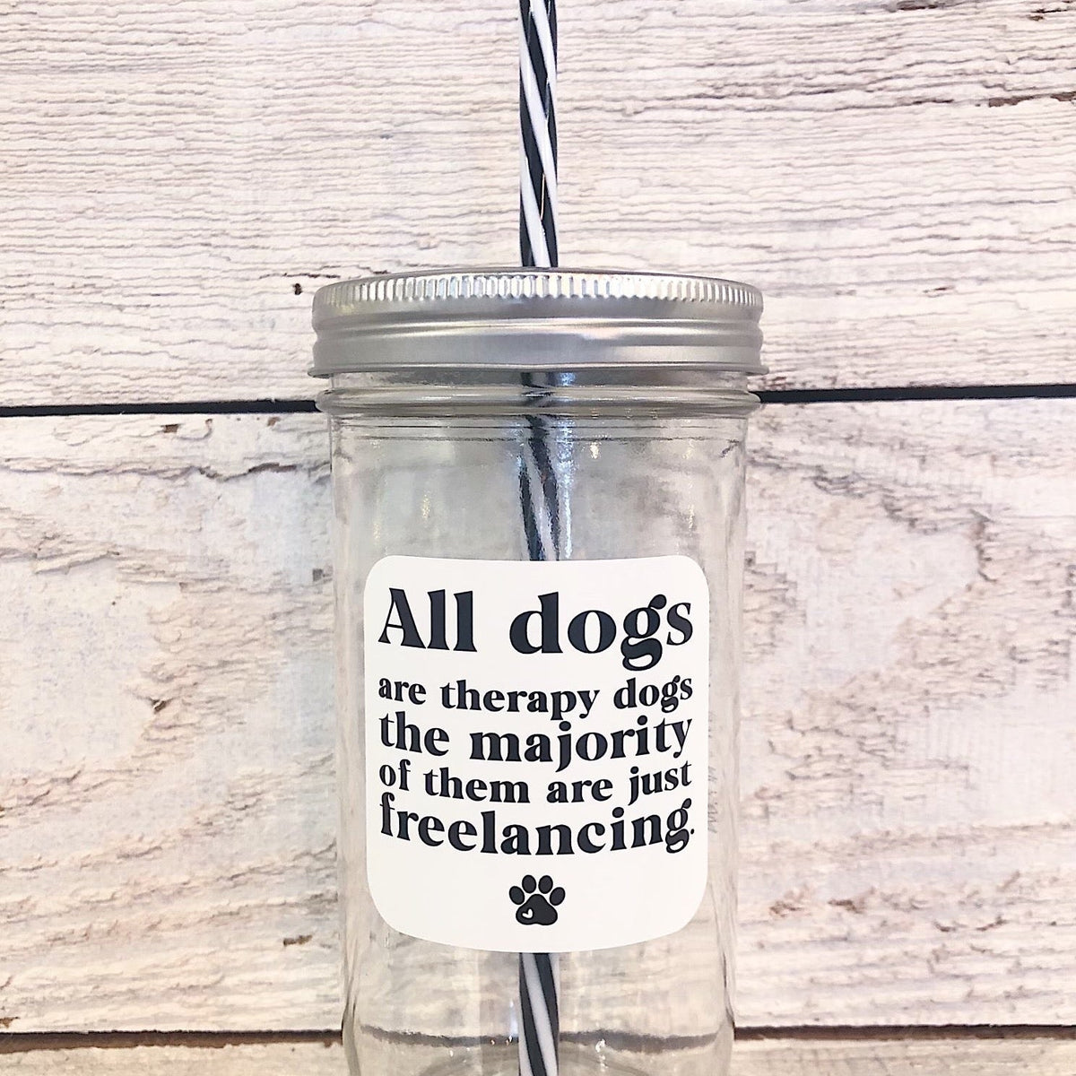 A mason jar tumbler that says “All dogs are therapy dogs the majority of them are just freelancing.” In black letters on a white background with a small black paw print at the bottom.