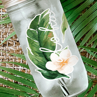 Close up of a mason jar that has a tropical leaf, white hibiscus flower, and “aloha” written in script.