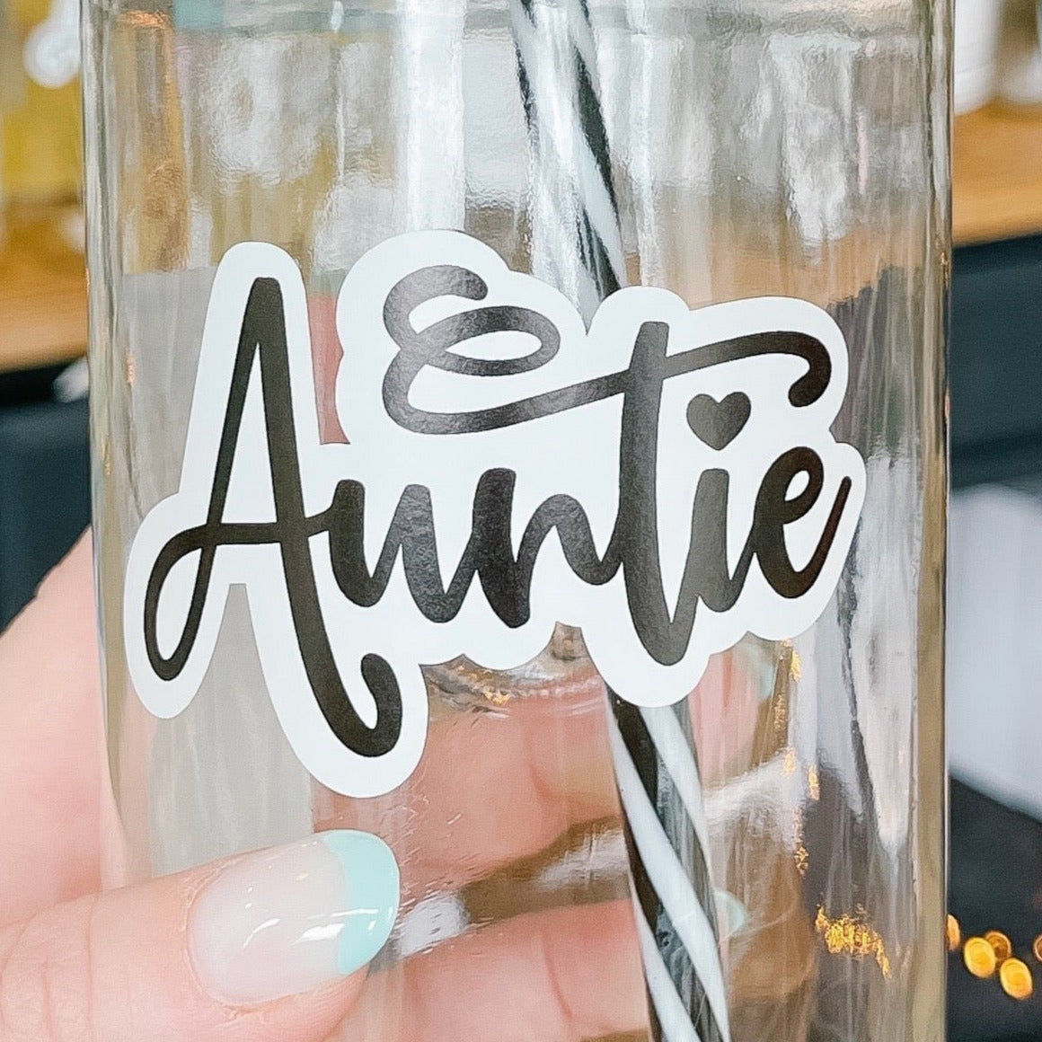 Close up of a hand holding a mason jar tumbler that says “Auntie” in script with the I dotted with a heart.