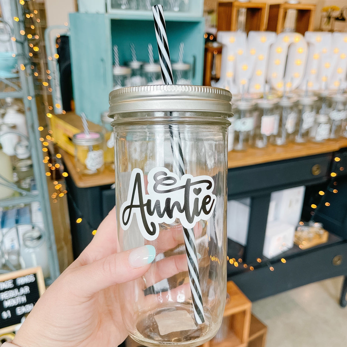 Hand holding a mason jar tumbler that says “Auntie” in script with the I dotted with a heart.