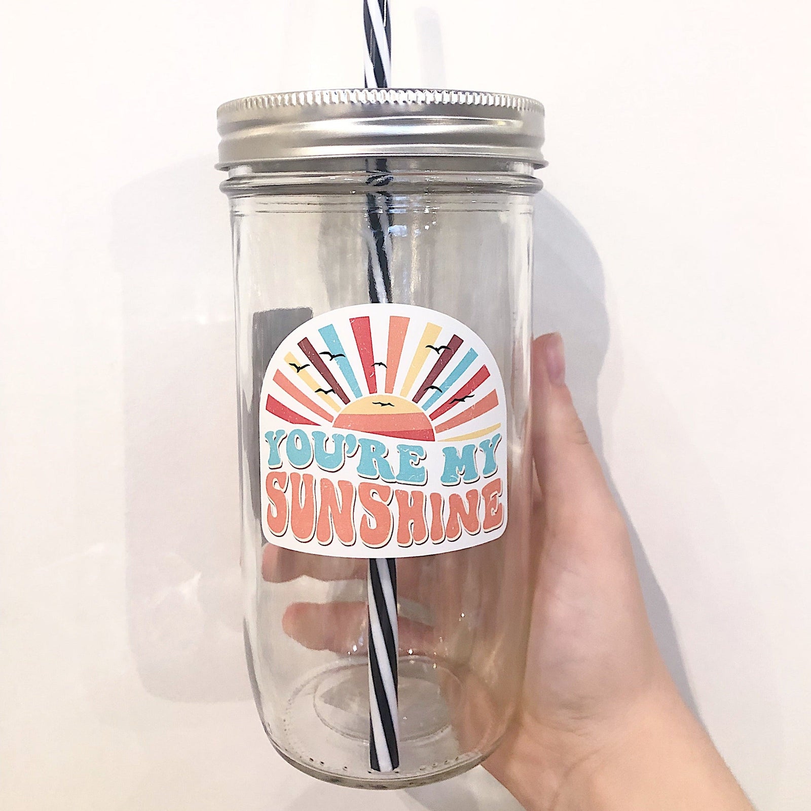 Hand holding a mason jar tumbler that has an image of a multicoloured sunset and says "you're my sunshine"