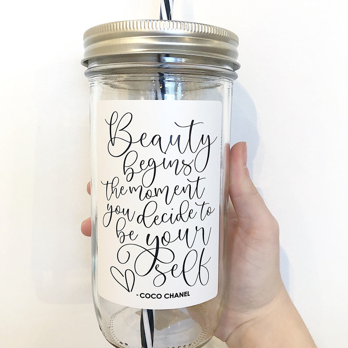 Hand holding a mason jar tumbler that says “Beauty begins the moment you decide to be your self -Coco Chanel” in black on a white background.