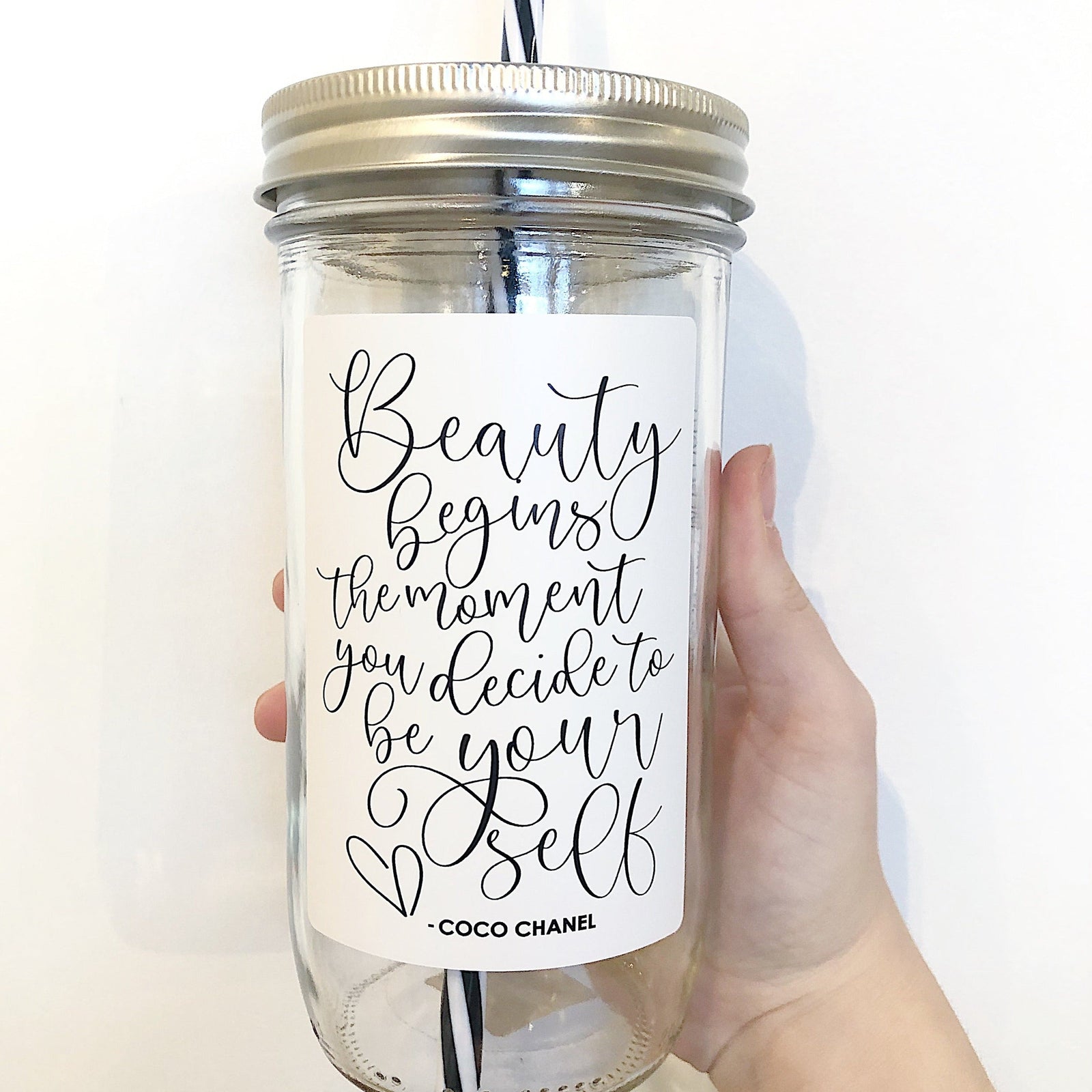 Hand holding a mason jar tumbler that says “Beauty begins the moment you decide to be your self -Coco Chanel” in black on a white background.