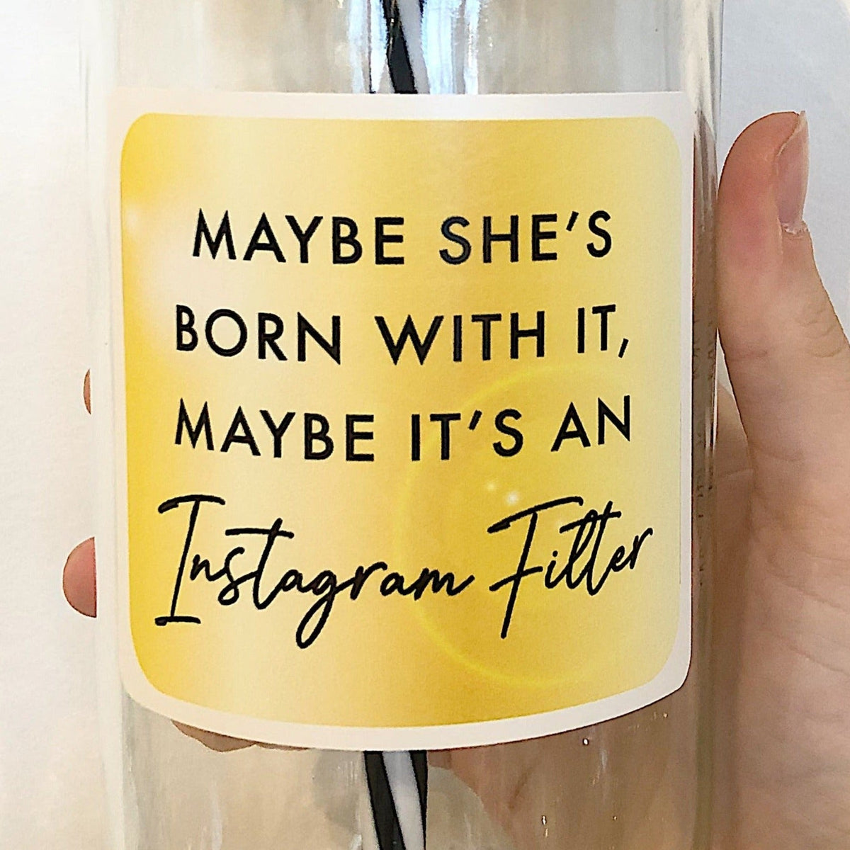 Close up of a hand holding a mason jar tumbler that has a square gold decal that reads “maybe she’s born with it, maybe it’s an instagram filter” in black lettering