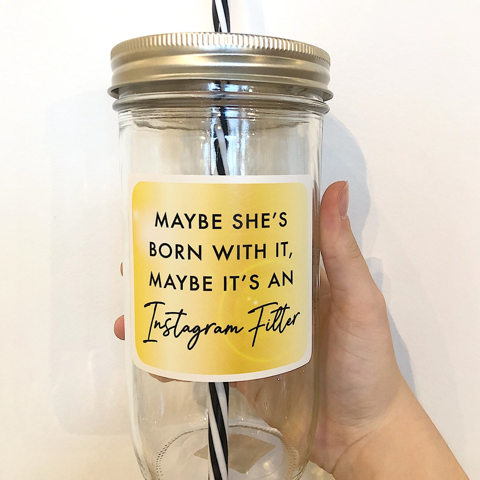 Hand holding a mason jar tumbler that has a square gold decal that reads “maybe she’s born with it, maybe it’s an instagram filter” in black lettering