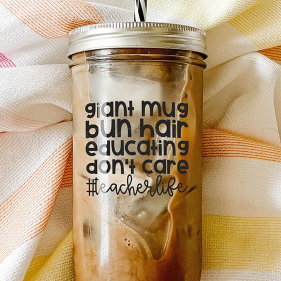 A #TeacherLife Mason Jar Tumbler with text that says "giant mug bun hair educating don't care". It has an iced coffee inside