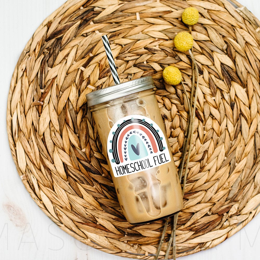Mason jar tumbler with iced coffee, laying over a fibre mat. Tumbler reads "Homeschool Fuel" with a rainbow image.