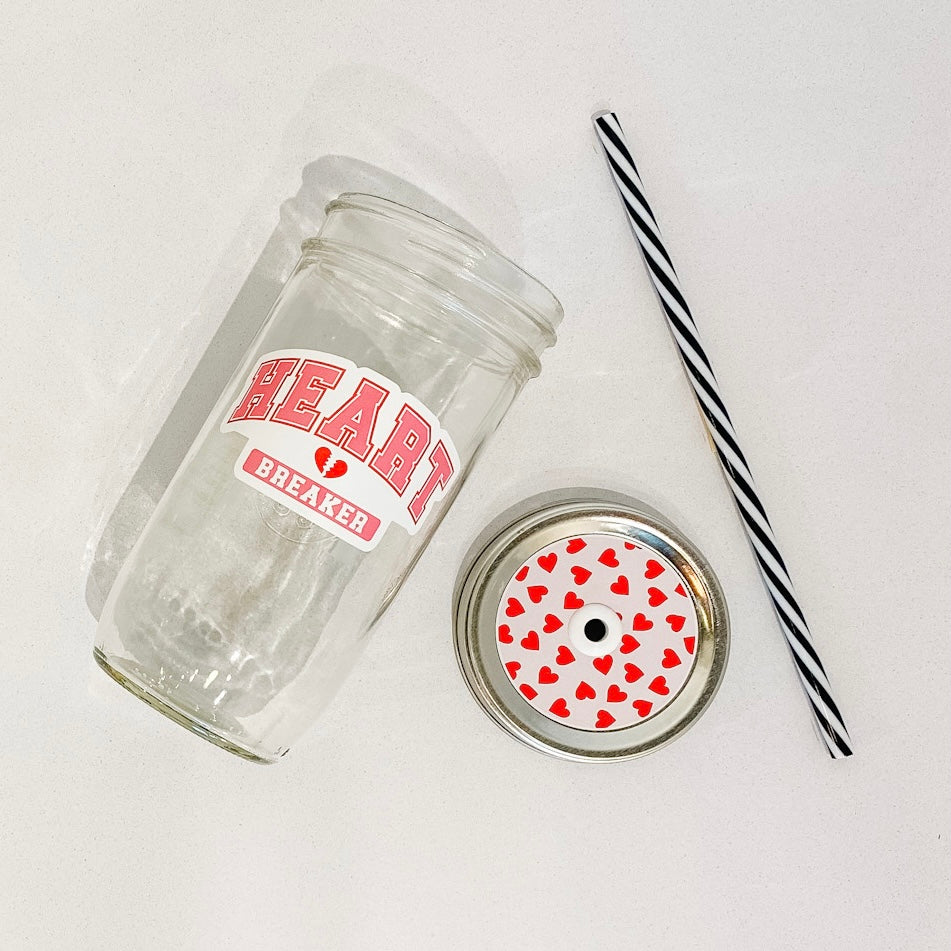 flat lay of the "HEART breaker" mason jar tumbler, a straw lid with with red hearts on a light pink background, and a black and white striped straw