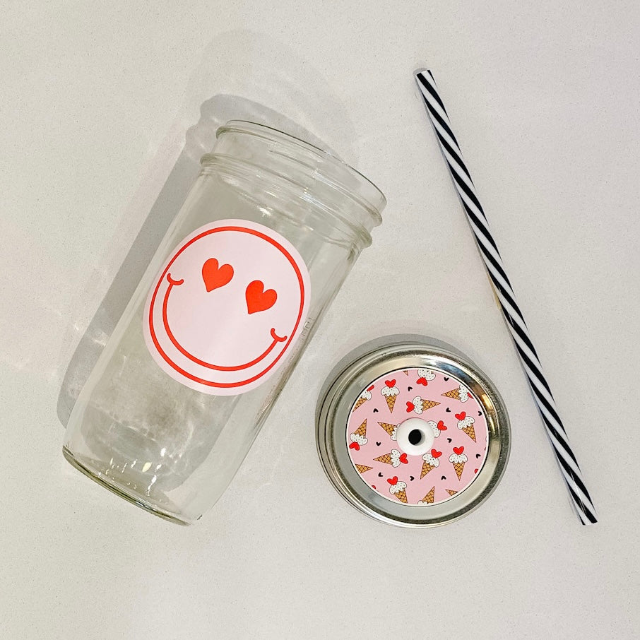 flat lay of the heart eyes smiley face mason jar tumbler, a straw lid with ice cream cones on a pink background, and a black and white striped straw