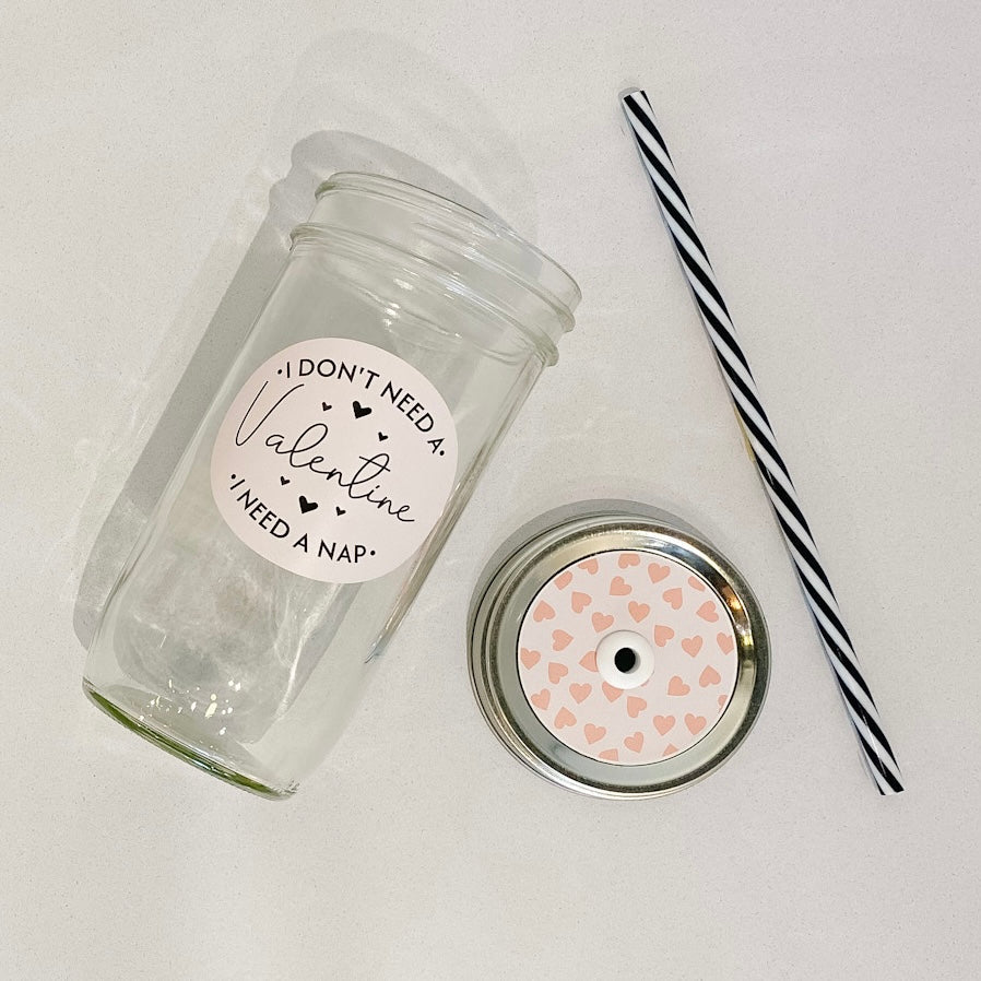 flat lay of the "I don't need a valentine" mason jar tumbler, a straw lid that has pink hearts on a light pink background, and a black and white striped straw