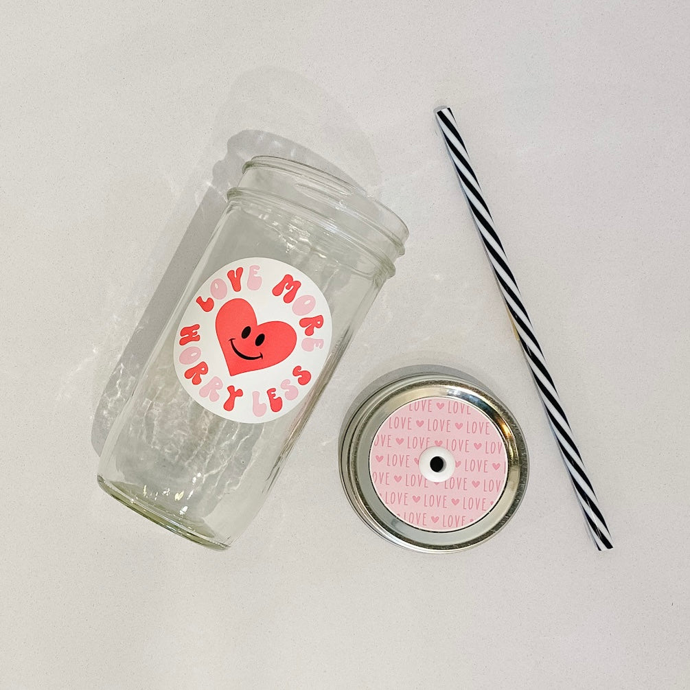 Flat lay of the "LOVE MORE WORRY LESS" mason jar tumbler, a straw lid that is light pink with dark pink "LOVE" written over and over, and a black and white striped straw