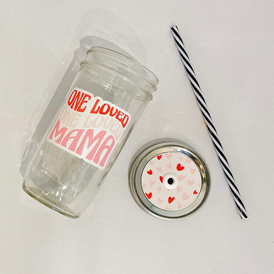 flat lay of the "one loved mama" mason jar tumbler, a straw lid with red and pink hearts on a light pink background, and a black and white striped straw