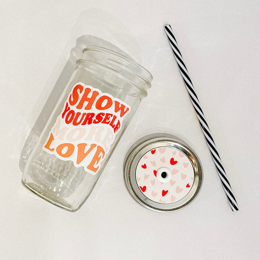 hand holding a mason jar tumbler that says "Show Yourself More Love" in a wavy retro font, in red, pink, and orange