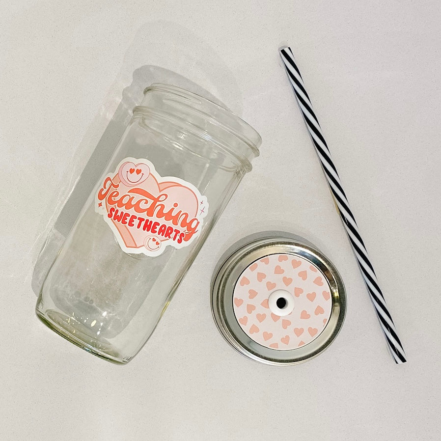 flat lay of the "Teaching Sweethearts" mason jar tumbler, a straw lid with pink hearts on a light pink background, and a black and white striped straw