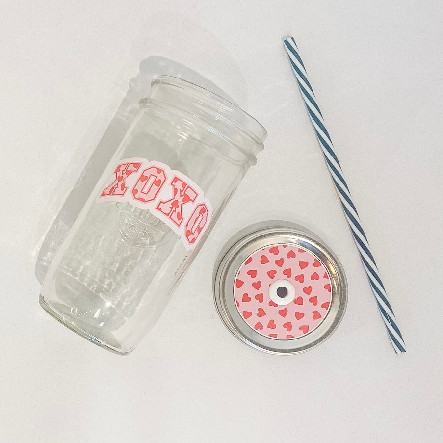 flat lay of a the "XOXO" mason jar tumbler, a straw lid with red hearts on a pink background, and a black and white striped straw