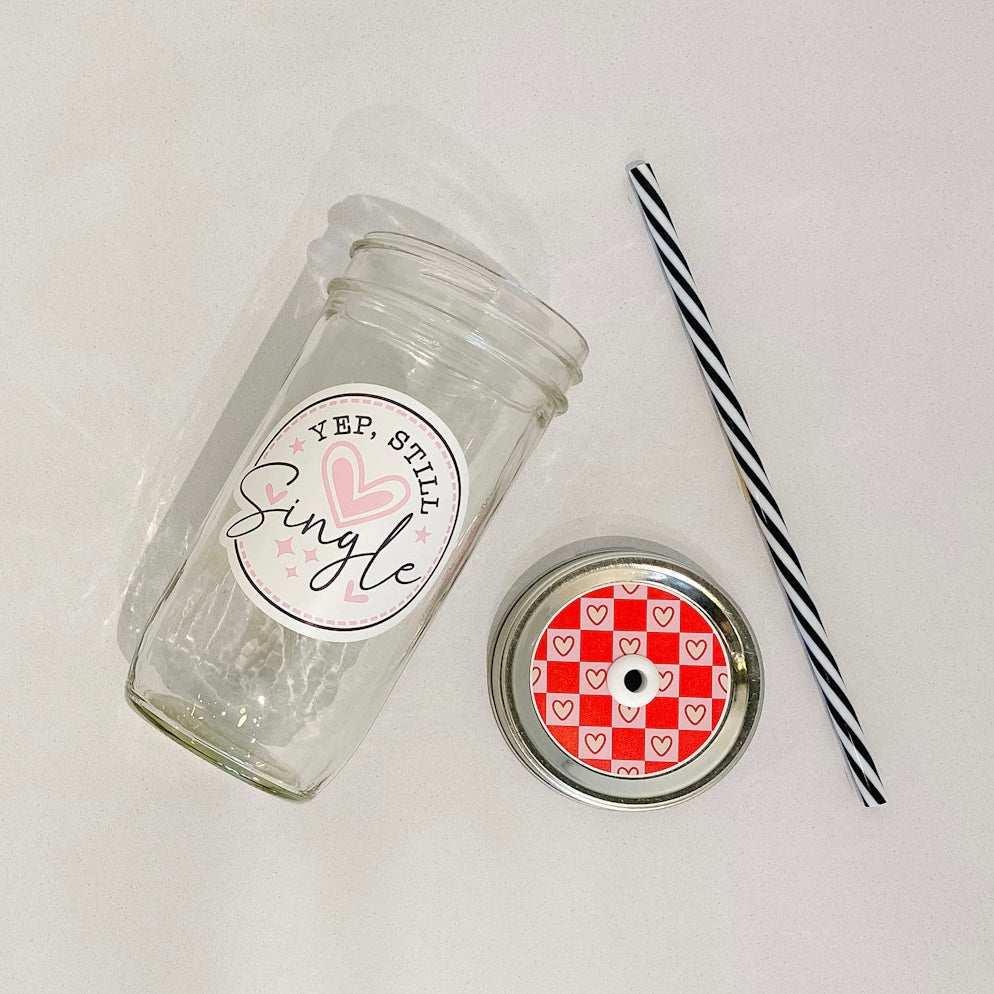 hand holding a mason jar tumbler that says "Yep, still single" on a white circular background with pink hearts
