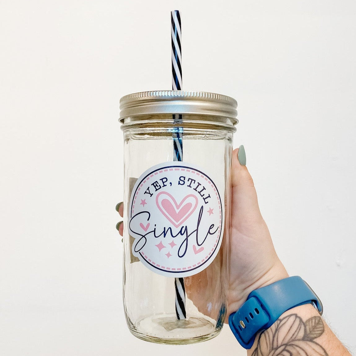 hand holding a mason jar tumbler that says "Yep, still single" on a white circular background with pink hearts