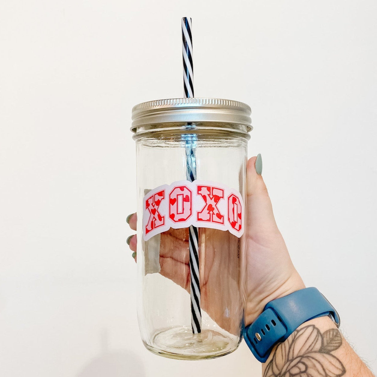 hand holding a mason jar tumbler that has "XOXO" in block letters with a red outline, the letters are filled with a light pink background and red hearts
