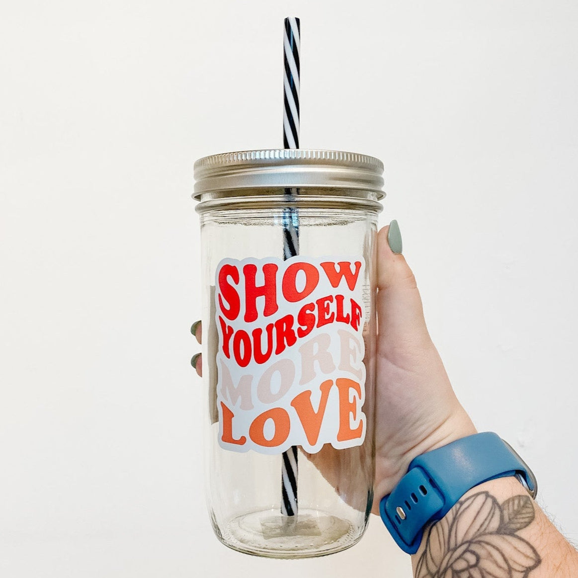 hand holding a mason jar tumbler that says "Show Yourself More Love" in a wavy retro font, in red, pink, and orange