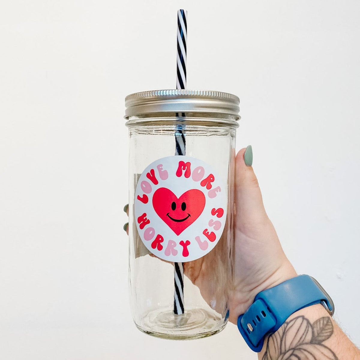 hand holding a mason jar tumbler that has a red heart with a black smiley face in the middle with the words "LOVE MORE WORRY LESS" in a circle around the heart. The lettering is bubbly and the letters alternate red and pink