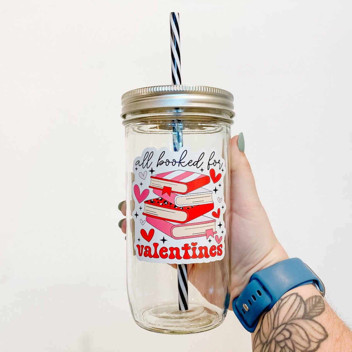 hand holding a mason jar tumbler that says "all booked for valentines" with a stack of books with red and pink covers, and red and pink hearts around