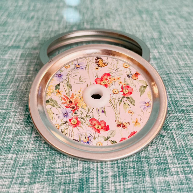 Mason jar patterned straw lid with a large assortment of flowers with a few butterflies on a light pink background.