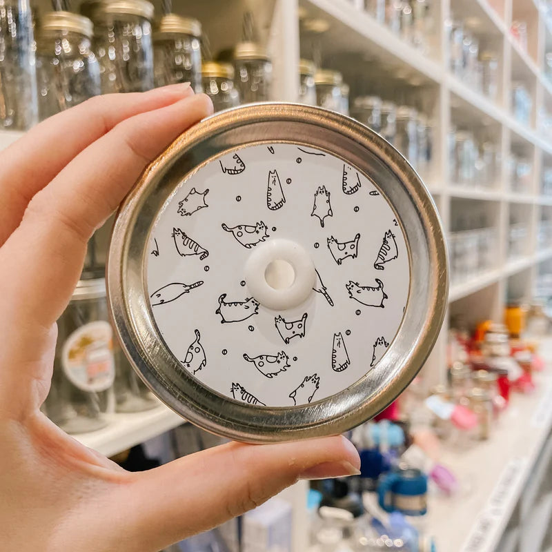 Hand holding mason jar patterned straw lid that has little black outlines of cats on white background