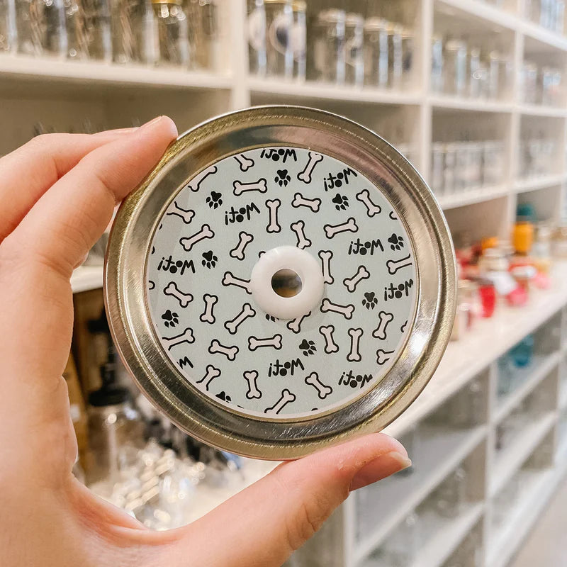 Hand holding mason jar patterned straw lid that has bones, paws and “woof!” On light blue background