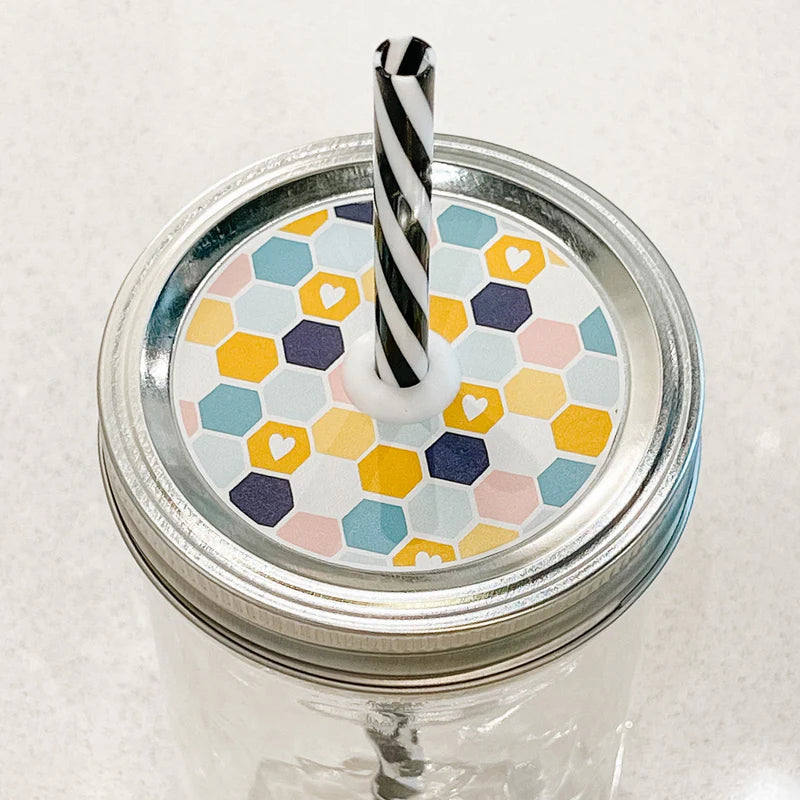 Hand holding mason jar patterned straw lid with multicoloured honey comb shapes and a few hearts.
