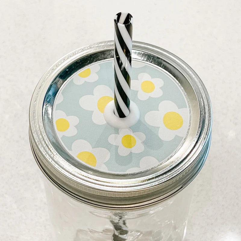 Hand holding mason jar patterned straw lid that has white and yellow daisies on light blue background