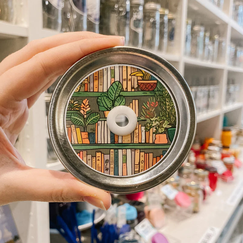 Hand holding mason jar patterned straw lid that has bookshelf and assorted plants