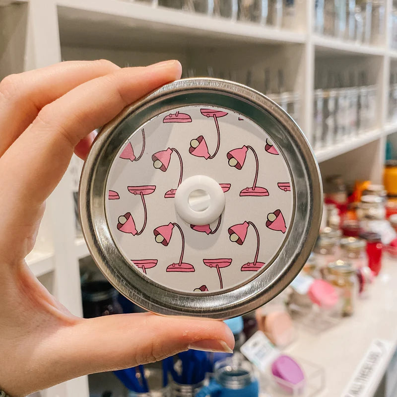 Hand holding mason jar patterned straw lid that has pink lamps on white background