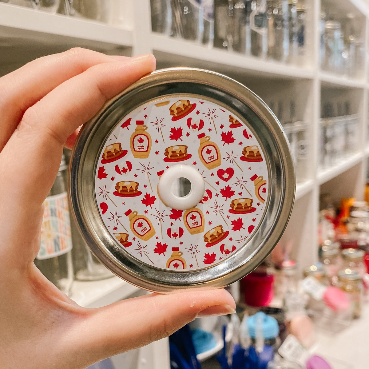 a straw lid that has a white background with maple leafs, canadian flags, pancake stacks, bottles of maple syrup, and fireworks all in a red, brown, and white colour scheme