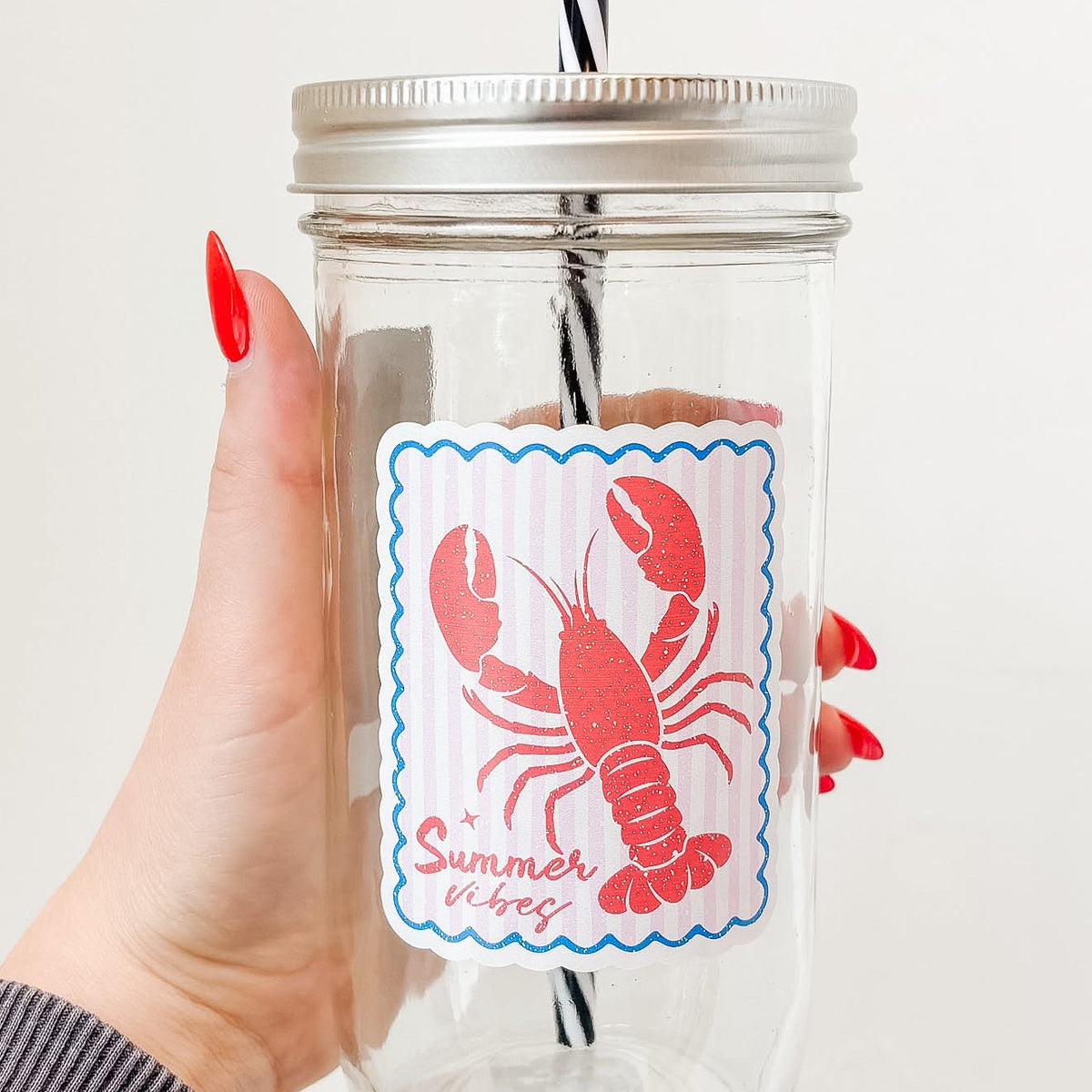 Hand holding mason jar tumbler up close that reads summer vibes in red ink with lobster design on pale pink and white striped background with blue border