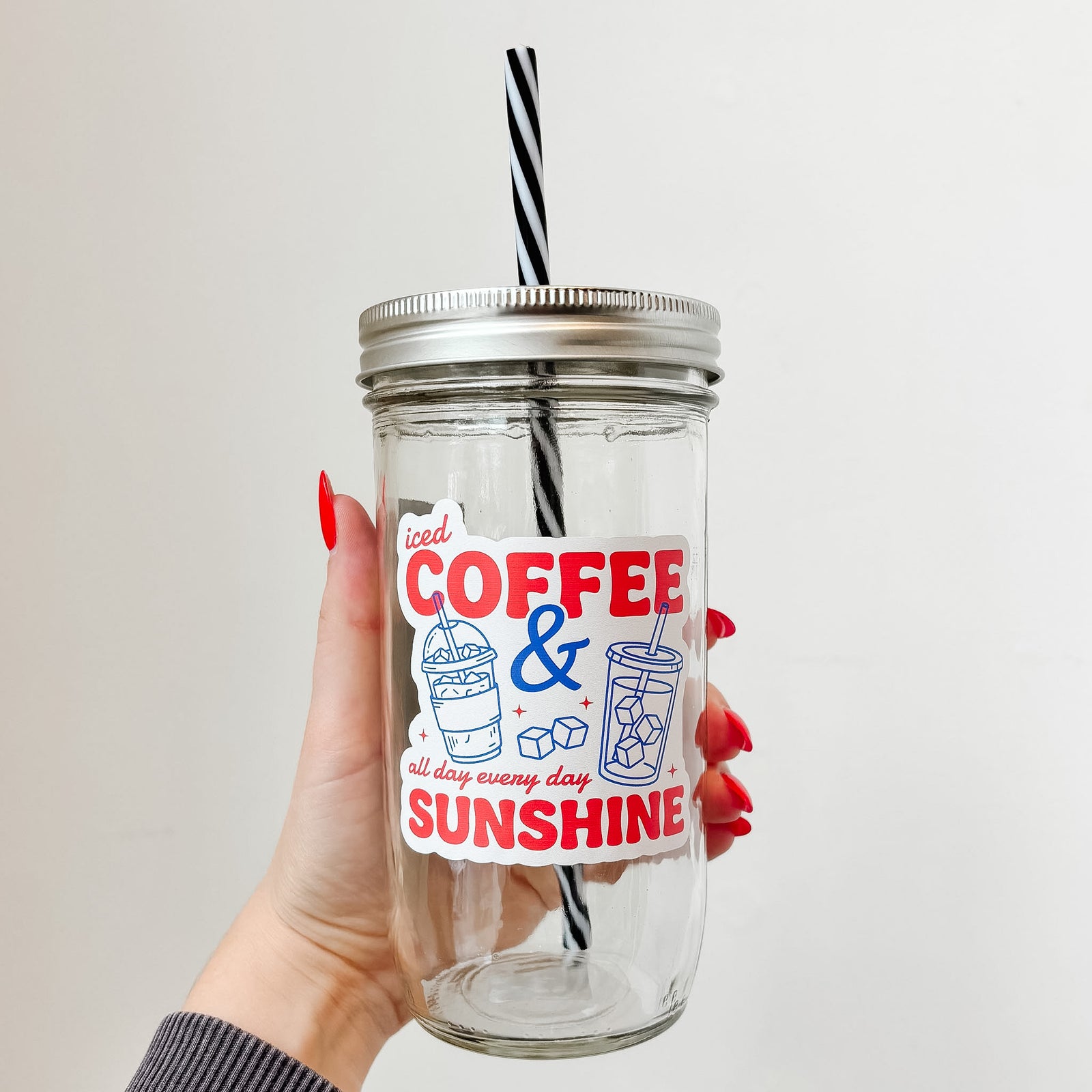 Hand holding mason jar tumbler that reads iced coffee & sunshine all day every day in red ink with blue iced drink designs on white background