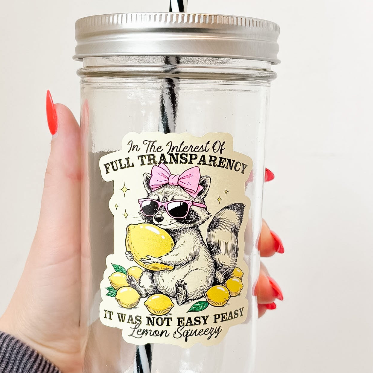 Hand holding mason jar tumbler that reads in the interest of full transparency it was not easy peasy lemon squeezy with a raccoon wearing pink sunglasses and bow holding a lemon and surrounded by lemons design on pale yellow background